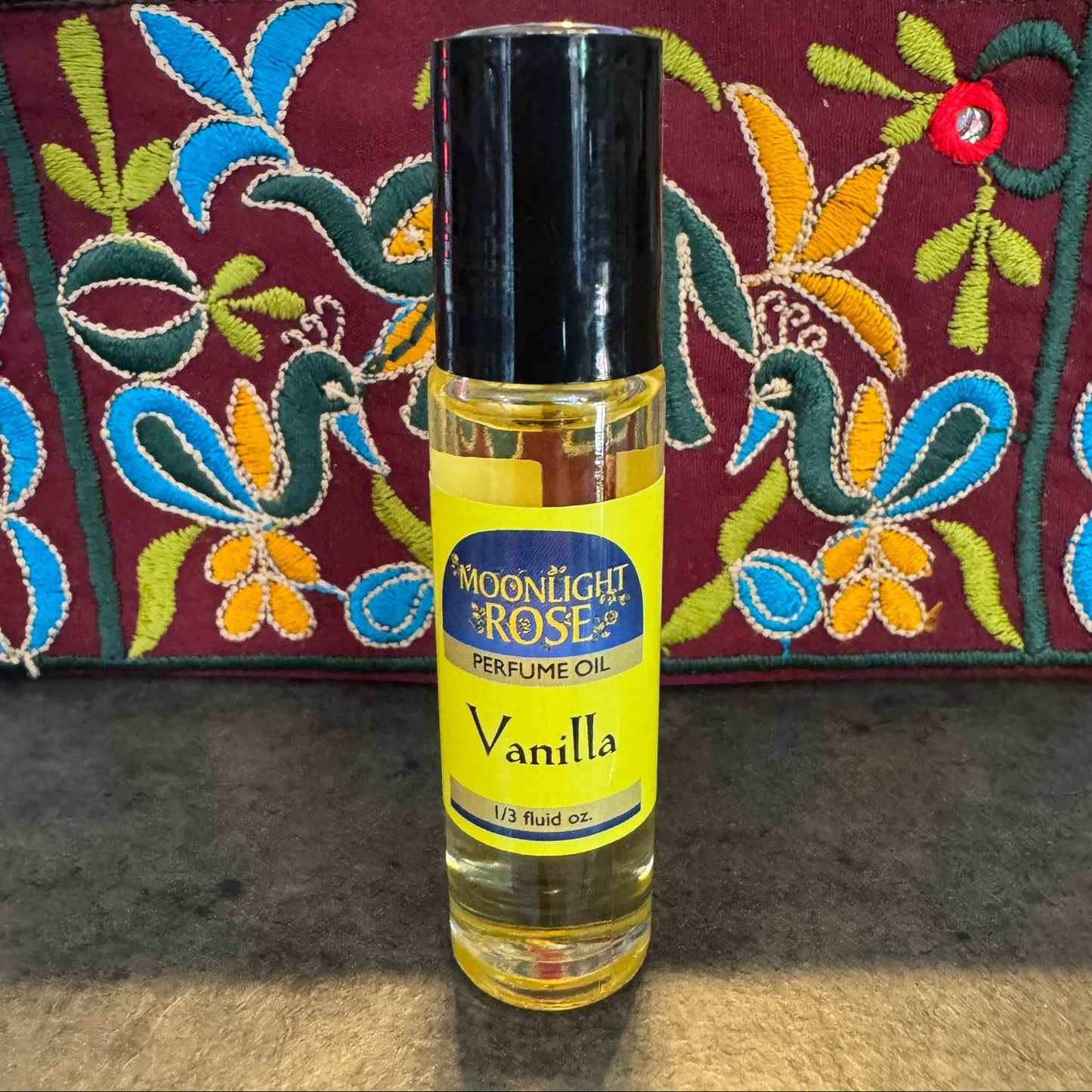 Perfume oil bottle labeled 'Vanilla' by 'Moonlight Rose' on a textured surface with a colorful embroidered fabric background.