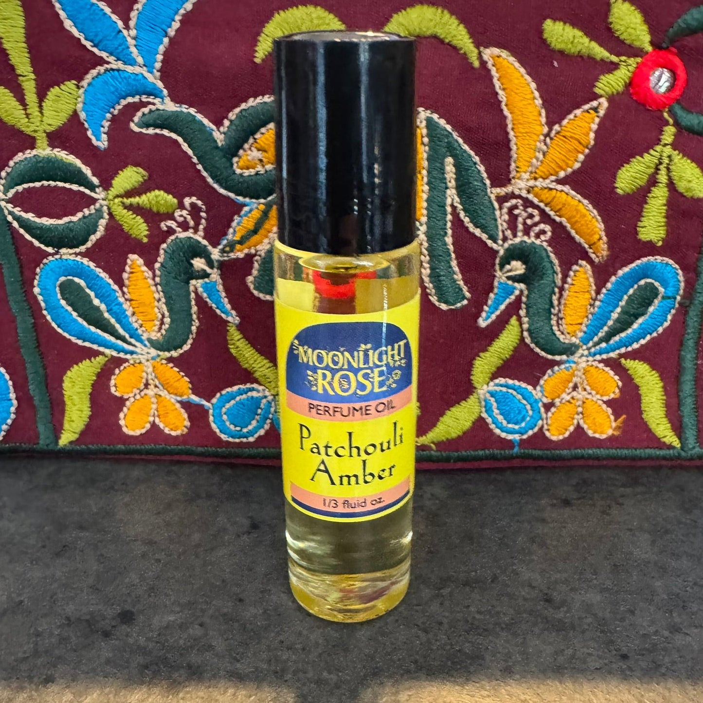 Bottle of Moonlight Rose Patchouli Amber perfume oil on a textured surface with an embroidered fabric background.