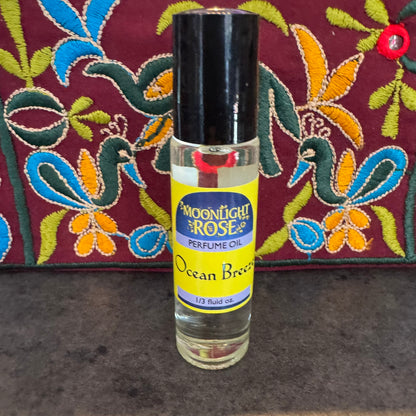 Perfume oil bottle labeled 'Moonlight Rose' with 'Ocean Breeze' on a textured surface with a colorful fabric background.